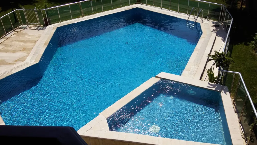 Bahçede yer alan korkuluklarla çevrili, entegre çocuk havuzlu geometrik açık yüzme havuzu.