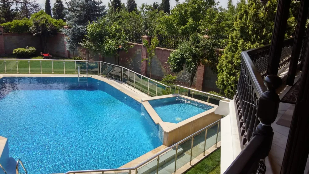 Ağaçlarla çevrili bahçede cam korkuluklu açık yüzme havuzu ve bitişiğindeki jakuzi.
